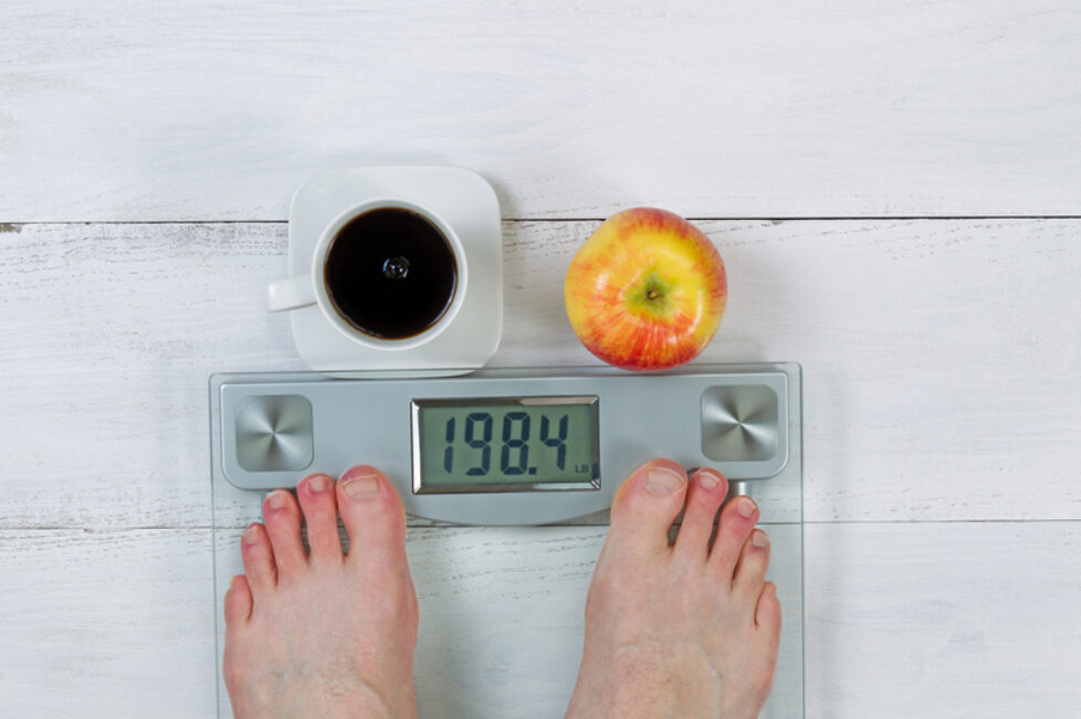 Photo of a weight scale, displaying body weight, with bare male feet and whole apple with black coffee in front on white wooden floor